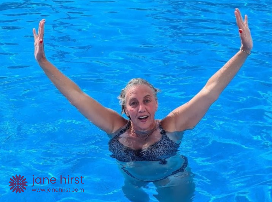 Lady in a bikini standing in a swimming pool. The water is shoulder high and her arms are raised up into the air