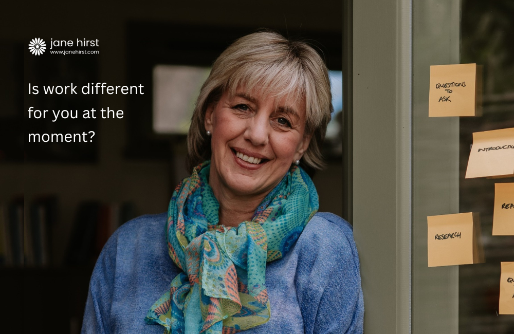 A lady leaning against an open door frame smiling at the camera. She has short blonde hair and is wearing a blue jumper with a brightly coloured scarf. The caption reads, 'Is work different for you at the moment?'